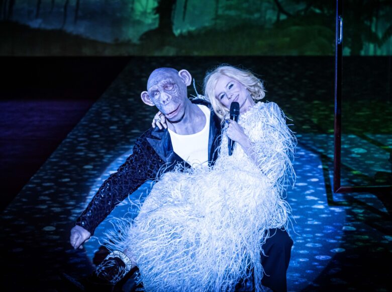 Scena teatralna. Mężczyzna w błyszczącej marynarce z maską małpy na twarzy trzyma na kolanach kobietę w białej sukni z piór. Kobieta w ręku trzyma mikrofon. Za nimi w półmroku fragment dekoracji przedstawiającej las nocą.