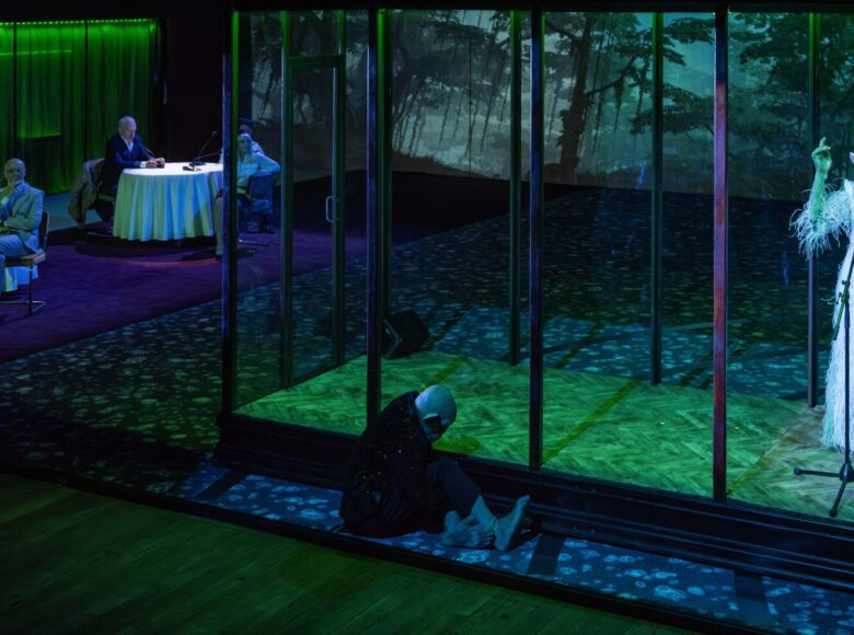 Przedstawienie teatralne. Na scenie dekoracja przedstawiająca wnętrze eleganckiej restauracji. Przy stolikach siedzi kilka osób. Na środku stoi wielka, szklana klatka. W stoi, śpiewając do mikrofonu, kobieta w wieczorowej sukni z maską małpy na twarzy. Przed klatka siedzi mężczyzna z maską małpy na twarzy.