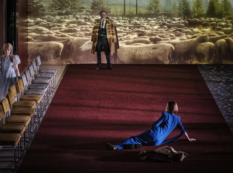 Na scenie leży czerwony dywan, wzdłuż niego stoi rząd krzeseł. W tle na ścianie powiększona fotografia stada owiec na pastwisku. Na pierwszym planie na dywanie siedzi kobieta. Za oparciami krzeseł stoi dziewczynka, a pod tylną ścianą stoi mężczyzna. Wszyscy są elegancko, odświętnie ubrani.