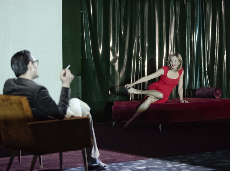 Scena w teatrze. Scenografia przedstawiająca pokój. Na łóżku siedzi kobieta w czerwonej, eleganckiej sukience. Naprzeciwko niej w fotelu siedzi mężczyzna i pali papierosa. Mężczyzna ubrany jest w ciemną marynarkę, jasne spodnie i sportowe buty.