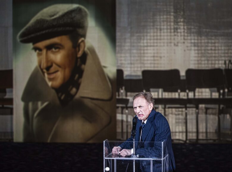 Na scenie, przy mównicy stoi starszy mężczyzna w eleganckiej marynarce. Za jego plecami na ścianie widzimy duży portret młodego mężczyzny w kaszkiecie i płaszczu, a obok powiększoną fotografię krzeseł stojących w rzędzie.