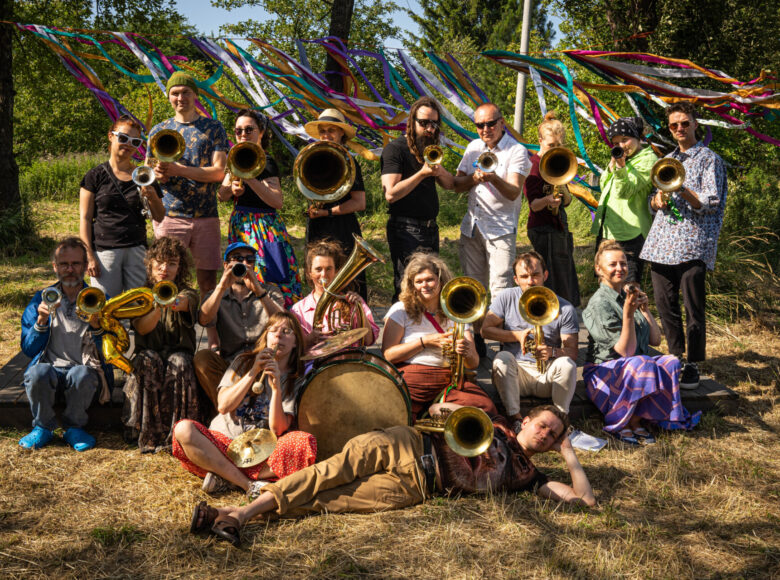 Słoneczny, letni dzień. Do zdjęcia na tle drzew i trawy pozuje grupa kolorowo ubranych młodych ludzi z muzycznymi instrumentami dętymi.