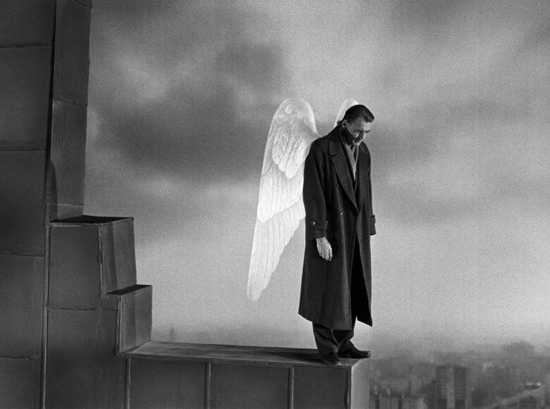 Kadr z filmu. Widok na miasto z lotu ptaka. Na pierwszym planie widzimy fragment muru, który wznosi się ponad dachami miasta. Na skraju muru stoi mężczyzna w eleganckim płaszczu i z anielskimi skrzydłami u ramion. Mężczyzna patrzy w dół.