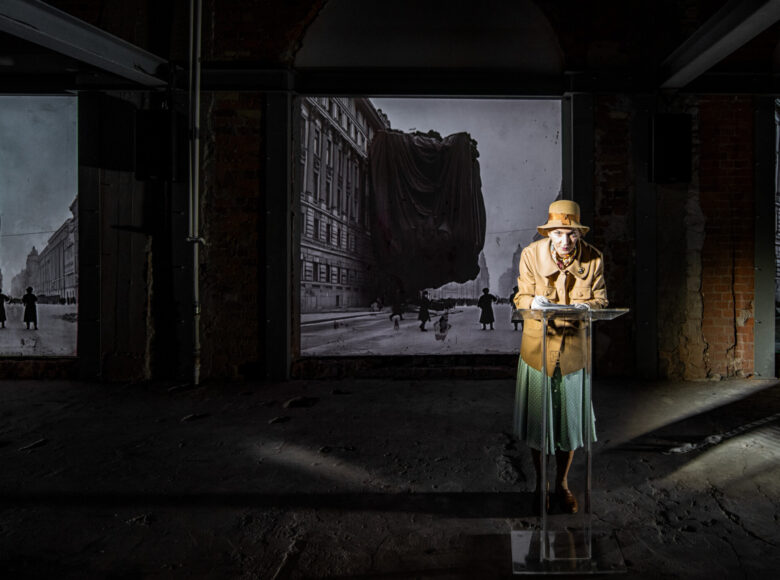 Na ceglanym murze wisi powiększona stara fotografia przedstawiająca kilka osób przed miejską kamienicą. Przed fotografią stoi mównica, przy mównicy kobieta w kapeluszu, żakiecie i spódnicy.