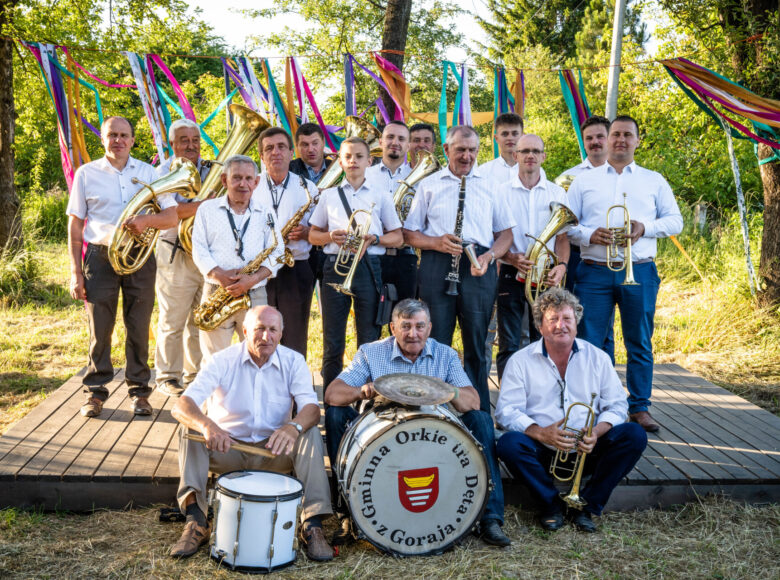 Słoneczny, letni dzień. Grupa elegancko ubranych mężczyzn z instrumentami muzycznymi pozuje do zdjęcia na tle drzew i trawy. Jeden z meżczyzn trzyma bęben z napisem: "Gminna Orkiestra Dęta z Goraja".