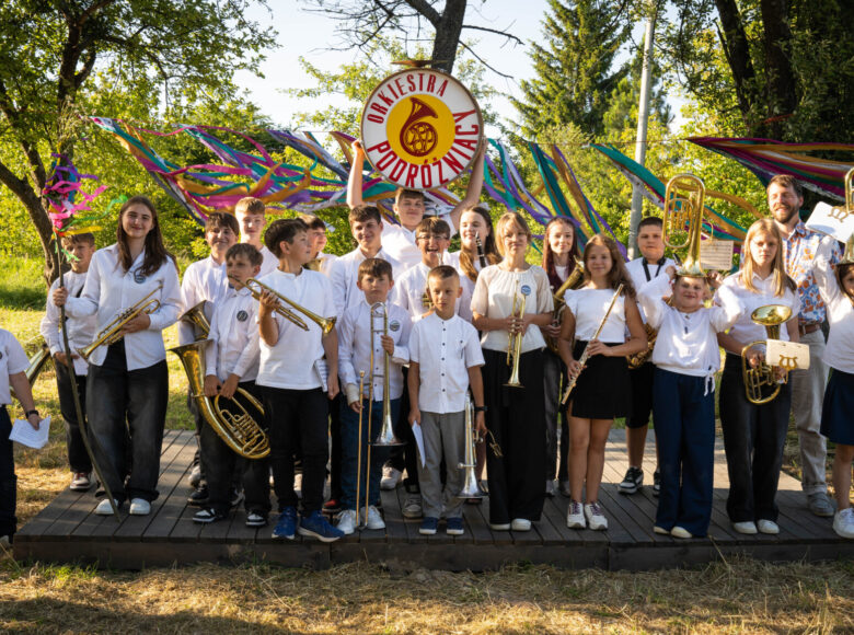 Słoneczny, letni dzień. Grupa elegancko ubranych dzieci z instrumentami muzycznymi pozuje do zdjęcia na tle drzew i trawy. Jedno z dzieci trzyma w rękach planszę z napisem: "Orkiestra Podróżniacy".
