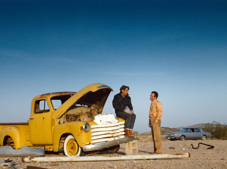Kadr z filmu. Na piaszczystym pustkowiu na tle niebieskiego nieba stoi stary, żółty samochód z paką. Na przedniej masce samochodu siedzi mężczyzna. Obok niego stoi drugi mężczyzna. Rozmawiają ze sobą. W tle zaparkowany samochód osobowy.