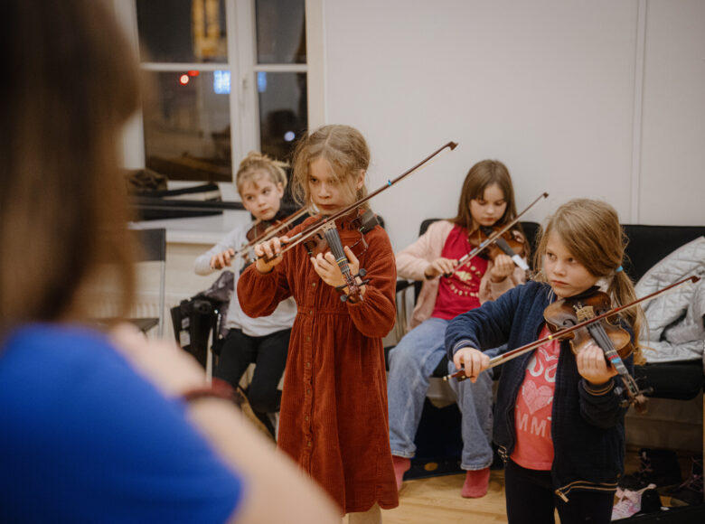 W sali grupka kilkuletnich dziewczynek grających na skrzypcach. Przed nimi stoi kobieta.