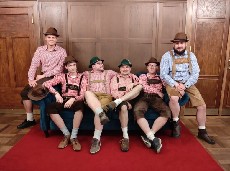 Grupa mężczyzn w bawarskich strojach pozuje do zdjęcia siedząc na sofie w pomieszczeniu.