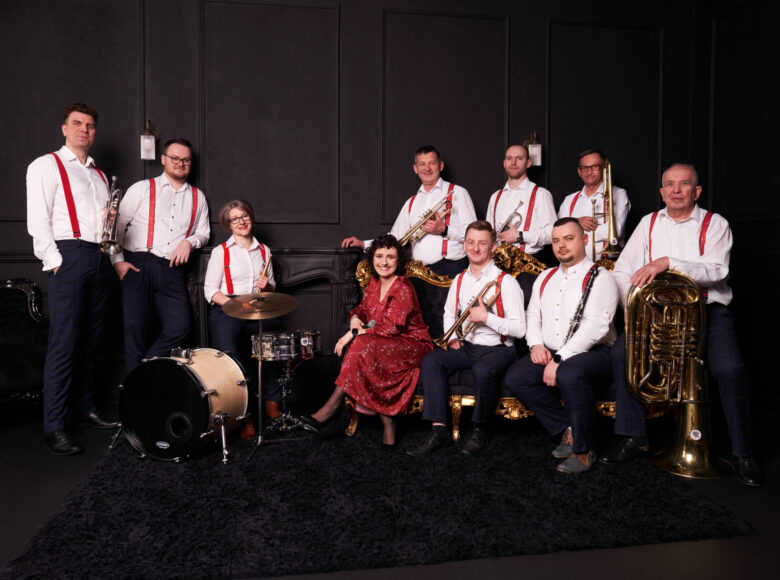 Grupa elegancko ubranych muzyków z instrumentami muzycznymi pozuje do zdjęcia na tle ciemnej ściany w pomieszczeniu.