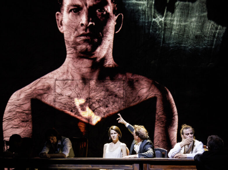 Spektakl teatralny. Przy stole siedzi kilka osób. Za nimi w tle ekran, na którym wyświetlana jest fotografia, przedstawiająca zbliżenie na mężczyznę z nagim torsem. Mężczyzna ten trzyma w dłoniach płonącą książkę.