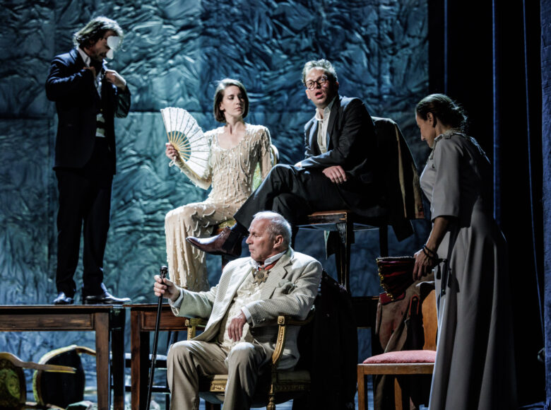 Spektakl teatralny. Dwa stoły. Obok stołów i na stołach stoją krzesła. Na krzesłach tych siedzi kilka osób w odświętnych strojach. Oprócz tego widzimy mężczyznę stojącego na jednym ze stołów oraz kobietę stojącą przy krześle przed stołami.