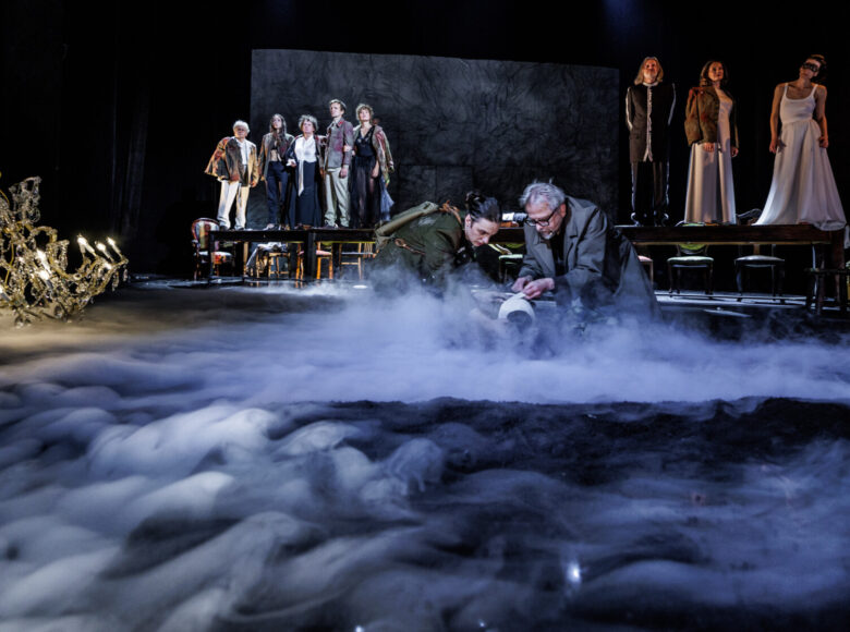 Spektakl teatralny. Na ziemi siedzi w kuckach dwóch mężczyzn, jeden z nich w mundurze żołnierza. Przed nimi snuje się dym. W tle duży stół, na którym stoją dwie grupy osób.