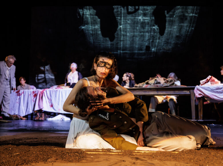 Spektakl teatralny. Na pierwszym planie siedząca na ziemi kobieta, podtrzymująca leżącego na jej kolanach mężczyznę w mundurze żołnierskim. Za nimi w tle duży stół zarzucony białymi obrusami, poplamionymi na czerwono. Przy stole siedzi grupa osób.