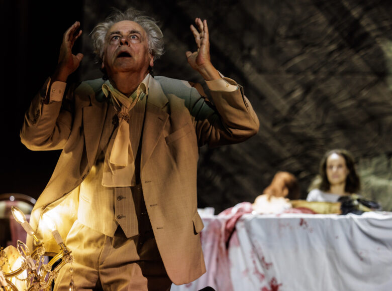 Spektakl teatralny. Na pierwszym planie mężczyzna ubrany w dziewiętnastowieczny garnitur wznosi do góry wzrok i ręce. Za nim stół. Na stół rzucony bezwładnie biały obrus poplamiony czerwoną substancją. Przy stole siedzi kobieta.