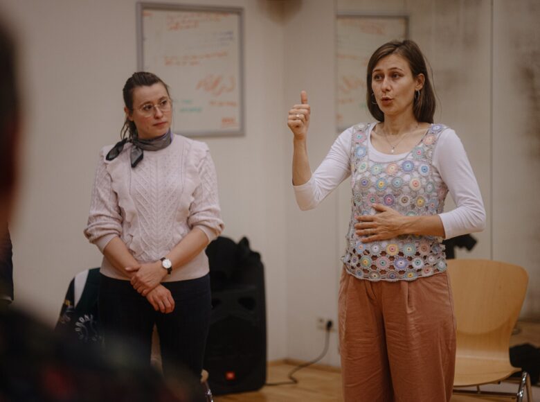 W pomieszczeniu stoją obok siebie dwie młode kobiety. Jedna pokazuje jak oddychać przeponą, druga na nią patrzy.