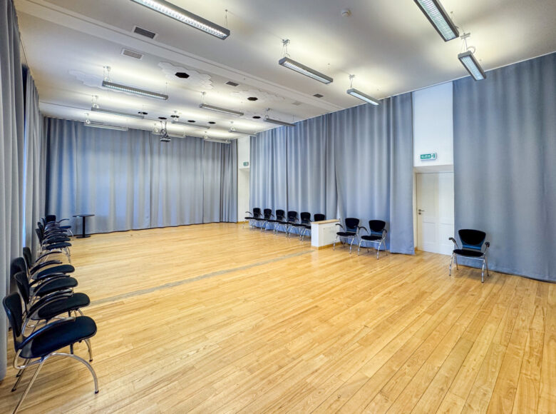 Wnętrze przestronnej, jasnej sali - tak zwanej "sali jasnej" w Centrum Kultury w Lublinie. W sali kilkanaście krzeseł ustawionych w rzędy pod ścianami. Ściany zasłonięte kotarami.