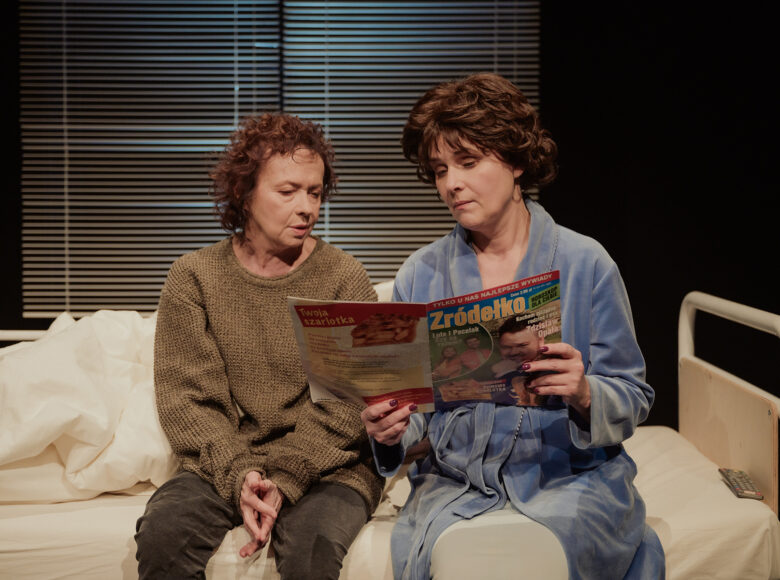 Spektakl teatralny. Scenografia przedstawiająca łóżko, stojące pod oknem. Na łóżku siedzą obok siebie dwie kobiety w średnim wieku i przeglądają gazetę. Jedna z kobiet ma na sobie szlafrok, druga spodnie i sweter.