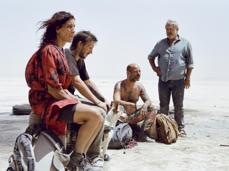 Na plaży nad morzem kobieta i trzech mężczyzn, siedzący na podróżnych tobołach. Wyglądają na zmęczonych i zgrzanych. Jest z nimi także pies.