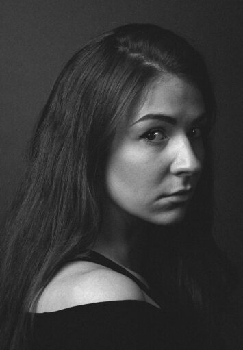 Czarno biały fotograficzny portret młodej kobiety na jednolitym, ciemnym tle.