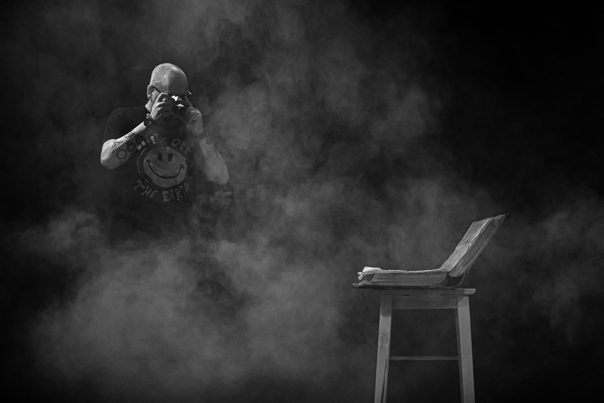 Czarno-biała fotografia, wykonana prawdopodobnie podczas spektaklu teatralnego, przedstawia na pierwszym planie krzesło na którym leży rozłożona wielka księga. Cały kadr fotografii spowija dym. Z tego dymu, w lewej części kadru wyłania się fotograf ubrany w czarną podkoszulkę z krótkim rękawem, unosi do oka w dłoniach aparat fotograficzny i robi tej księdze zdjęcia.