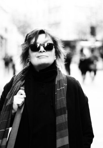 Fotografia czarno biała. Kobieta w średnim wieku ubrana w płaszcz, szalik i okulary słoneczne stoi na miejskiej ulicy, pozując do fotografii.