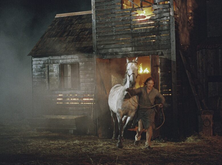 Kadr z filmu. Noc. Z płonącego drewnianego budynku wybiega mężczyzna, prowadząc konia.