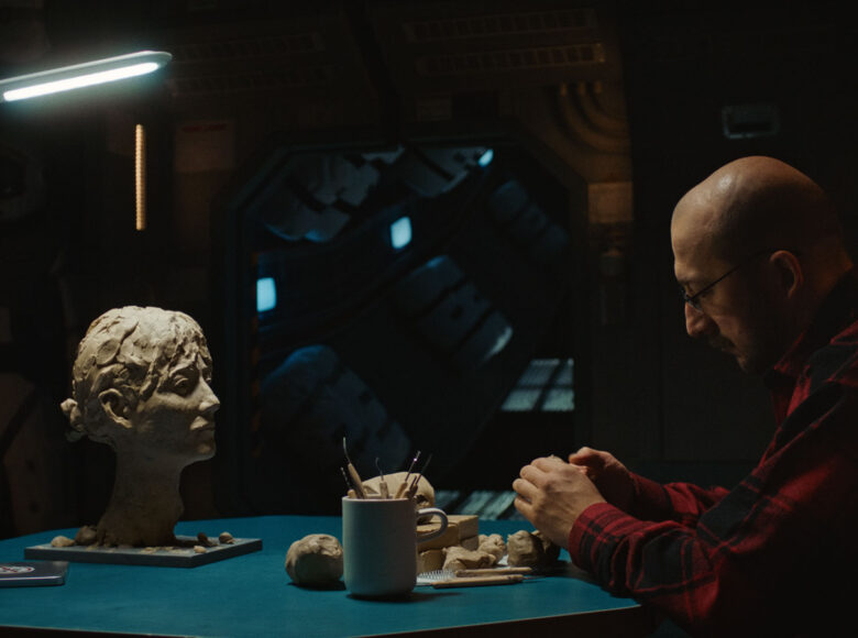 Ciemne pomieszczenie. Na pierwszym planie punktowo oświetlony stół. Przy stole siedzi mężczyzna i rzeźbi głowę kobiety. Na blacie stołu leżą kawałki gliny i narzędzia rzeźbiarskie.