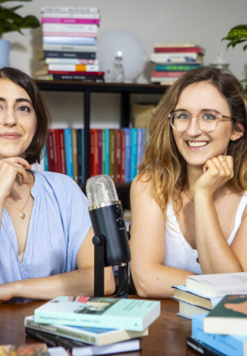 Przy stole zarzuconym książkami siedzą dwie młode kobiety. Za nimi półki z książkami i donice z zielonymi, wysokimi roślinami.