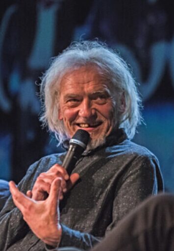 Zbliżenie na twarz i ręce siedzącego starego mężczyzny, który mówi coś do mikrofonu patrząc na kogoś, kogo nie widać na fotografii. Mężczyzna nie ma przedniego zęba. Z tyłu niedookreślone, niebiesko czarne tło.