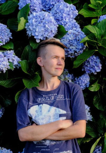 Fotograficzny portret młodej kobiety w niebieskiej bluzce, stojącej na tle kwitnących na niebiesko hortensji.