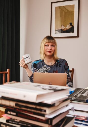 Młoda kobieta siedzi przy stole. Przed nią na blacie stołu laptop i stosy książek. W ręku kobieta trzyma kubek z napisem "Wisława".