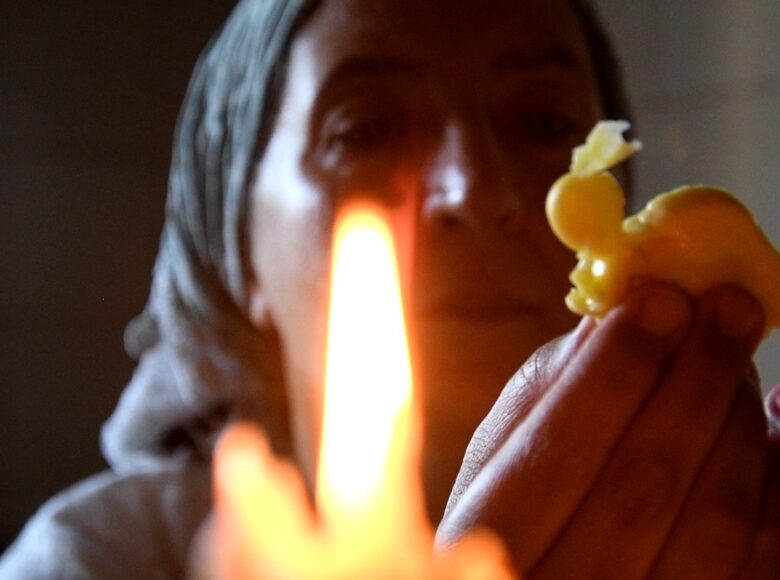 Kadr z filmu. Zbliżenie na twarz i dłonie wiejskiej kobiety w chustce. Kobieta patrzy na trzymany w dłoni kawałek stopionego wosku. Obok stoi płonąca świeczka.
