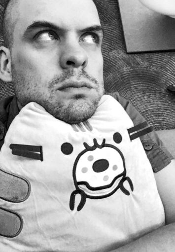 Czarno biały portret fotograficzny młodego mężczyzny. Mężczyzna siedzi i trzyma przed sobą poduszkę, na której opiera brodę.