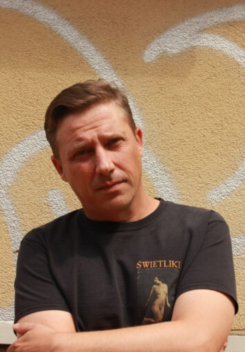 Fotograficzny portret mężczyzny w średnim wieku na tle żółtego muru, pokrytego napisami graffiti.