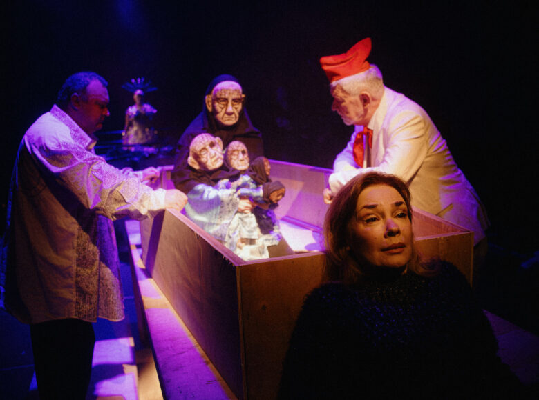 Spektakl teatralny teatru lalki i aktora. Na scenie na pierwszym planie kobieta pokazana od pasa w górę. Za nią trzy osoby odgrywające scenę za pomocą lalek teatralnych - dwóch mężczyzn i osoba zasłonięta maską na twarzy i peleryną.