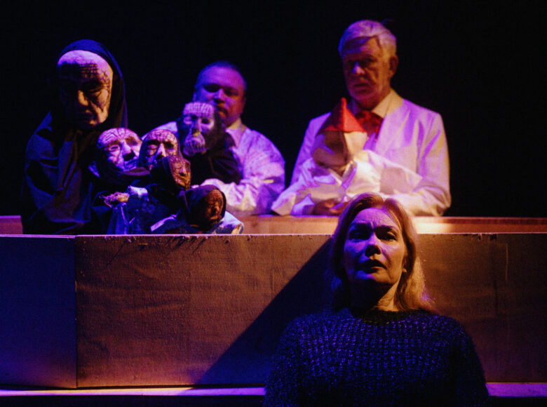 Spektakl teatralny teatru lalki i aktora. Na scenie na pierwszym planie kobieta pokazana od pasa w górę. Za nią trzy osoby odgrywające scenę za pomocą lalek teatralnych - dwóch mężczyzn i osoba zasłonięta maską na twarzy i peleryną.