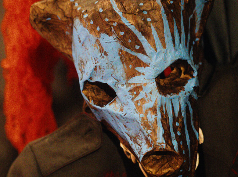 Spektakl teatralny. Zbiżenie na osobę z maską wilka na twarzy. Z tyłu niedookreślony, czerwony kształt.