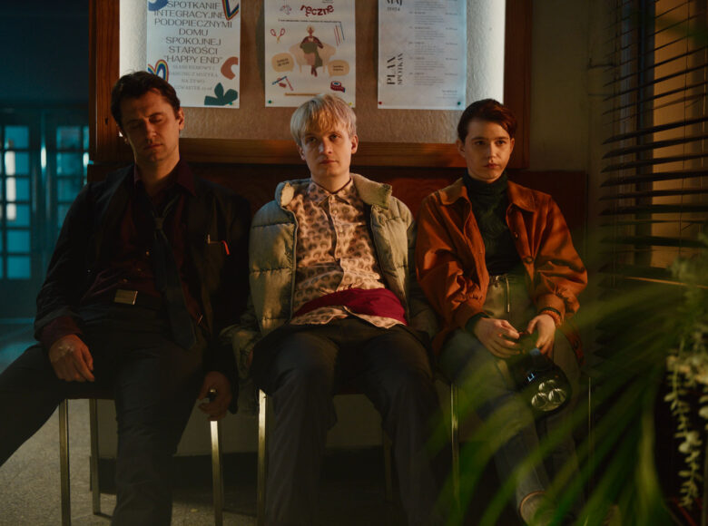 Kadr z filmu. W budynku użyteczności publicznej siedzą obok siebie na krzesłach dziewczyna i dwóch chłopaków.