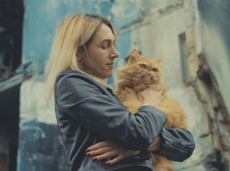 Kadr z filmu. Na pierwszym planie zbliżenie na dziewczynę, trzymającą na rękach kota. Za nią ruiny kamienicy, uszkodzonej przez wybuch.