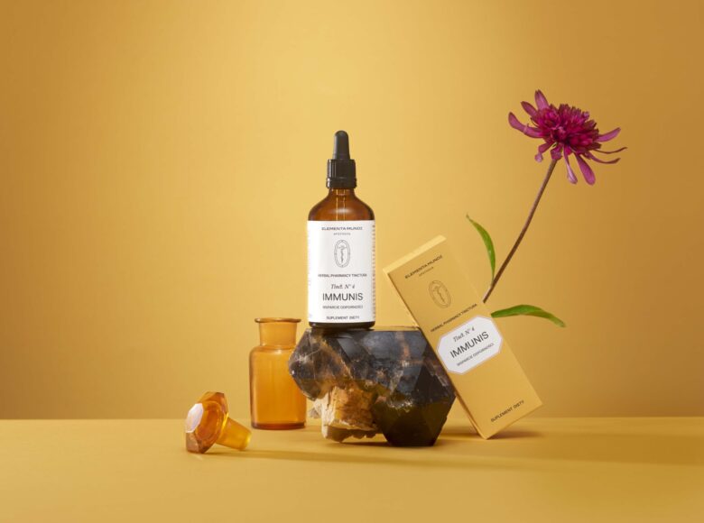 Na żółtym tle ustawione pudełeczko i flakoniki z kosmetykami oraz kwiat.