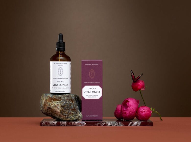 Na ciemnym tle ustawione pudełeczko i flakonik z kosmetykami oraz kilka główek kwiatów.