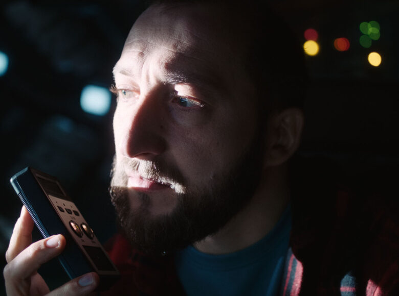 Kadr z filmu. Zbliżenie na twarz młodego mężczyzny z brodą. Mężczyzna trzyma w dłoni przy swoich ustach małe urządzenie elektroniczne. Za mężczyzną czarne tło z kilkoma punktami kolorowego światła.