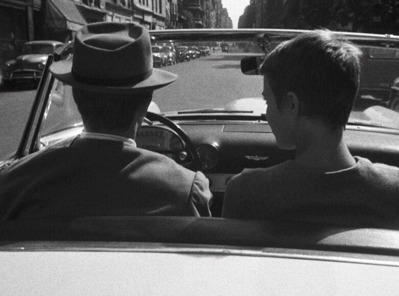 Kadr z filmu czarno biały. Zbliżenie na pokazanych od tyłu chłopaka i dziewczynę jadących samochodem. Chłopak siedzi za kierownicą, dziewczyna obok niego. Przed nimi rozciąga się widok miejskiej ulicy.