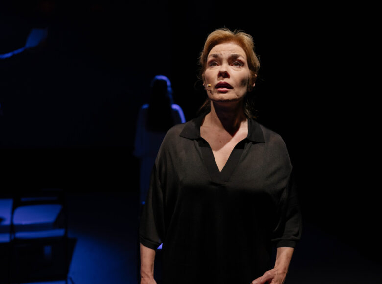 Spektakl teatralny. Na scenie na pierwszym planie stoi kobieta. Za nią w oddaleniu stoi druga kobieta. Z tyłu czarne tło.