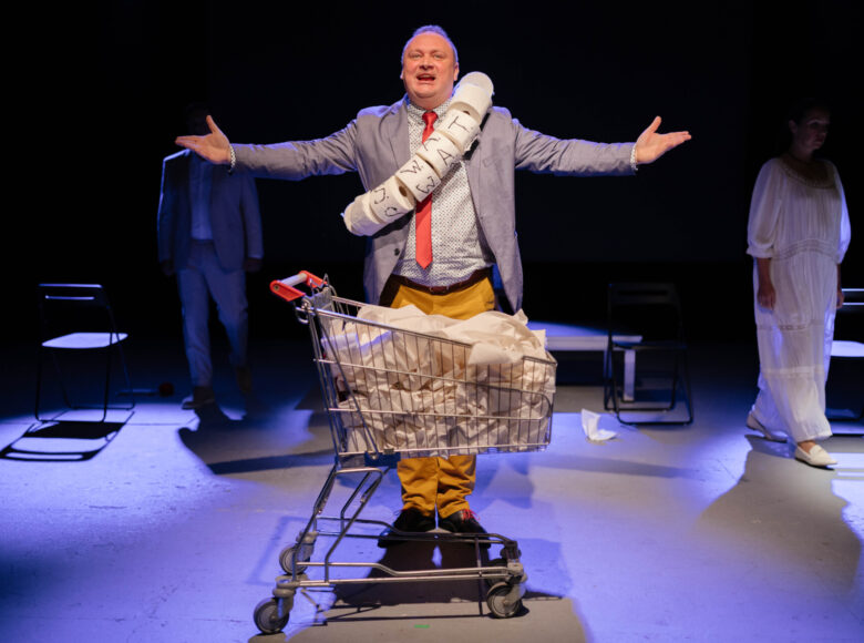 Przedstawienie teatralne. Na pierwszym planie stoi mężczyzna z rolkami papieru toaletowego zawieszonymi na szyi. Obok stoi wózek na zakupy. W tle drugi mężczyzna i kobieta, idący w stronę pierwszego planu. Z tyłu czarne tło.