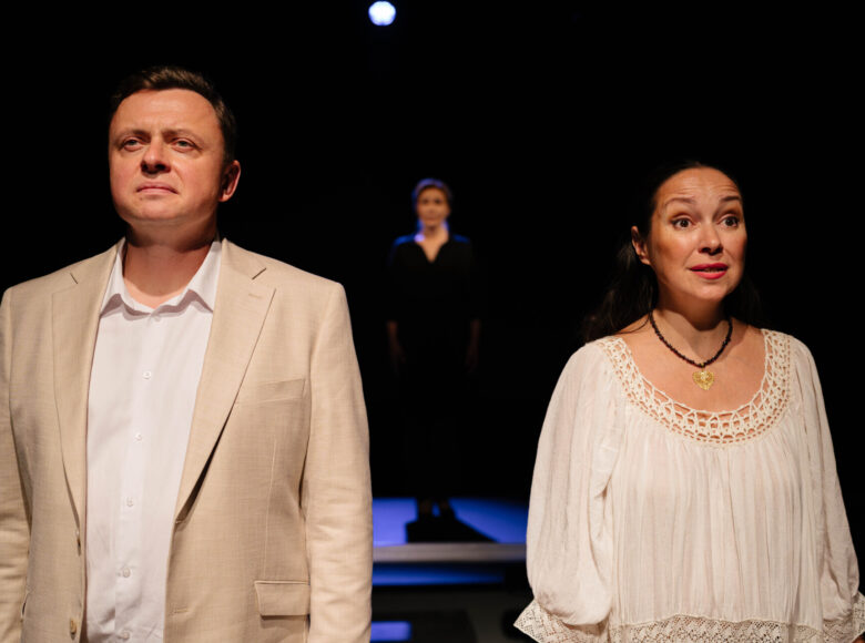 Spektakl teatralny. Na scenie stoją obok siebie kobieta i mężczyzna w białych ubraniach. Za nimi na dalszym planie kobieta ubrana na czarno. Z tyłu czarne tło.
