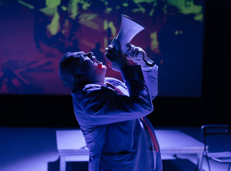 Spektakl teatralny. Na scenie mężczyzna krzyczacy do megafonu. Za nim w tle na ekranie projekcja multimedialna.