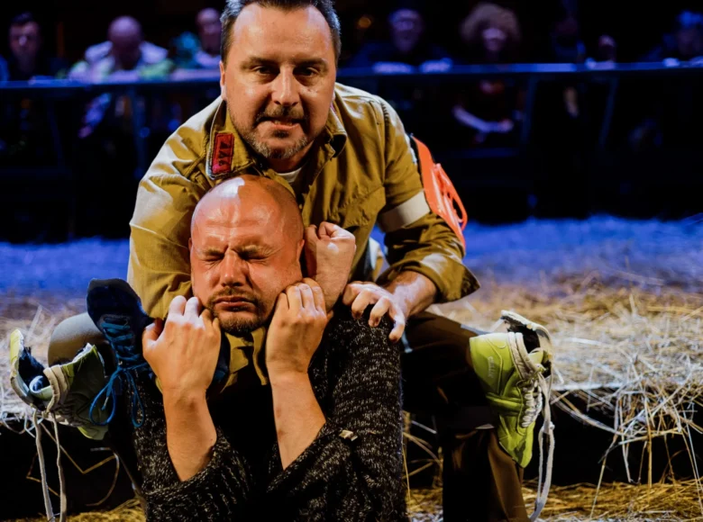 Spektakl teatralny. Zbliżenie na dwóch mężczyzn. Jeden z nich, ubrany w mundur, obejmuje ramieniem szyję drugiego jakby go dusił.