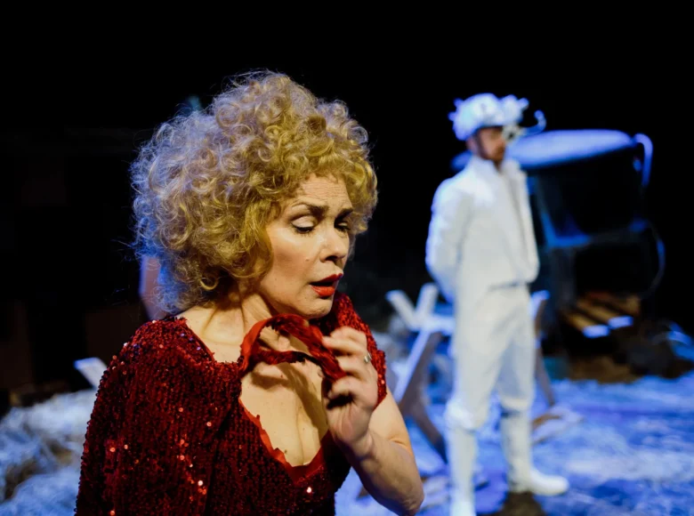 Spektakl teatralny. Na pierwszym planie stoi kobieta w czerwonej, błyszczącej sukni. W głębi stoi ubrany na biało mężczyzna. Za nimi fragmenty scenografii i ciemne, czarne tło.